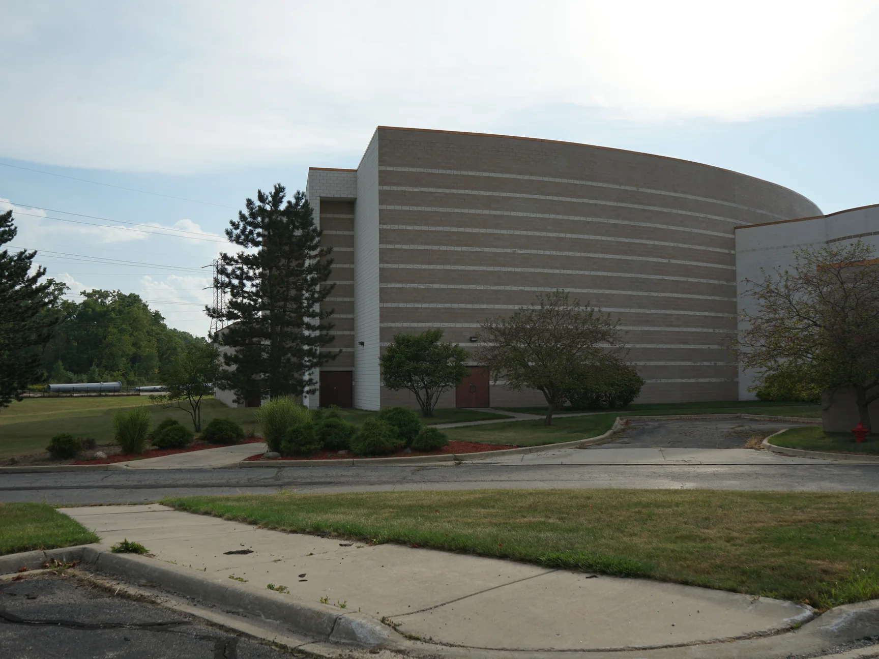 The building from the road in 2019, a tall curved block volume with light accent courses stepping across it, joined to a long low wing, with pines at the left and a parking drive and lawn across the foreground