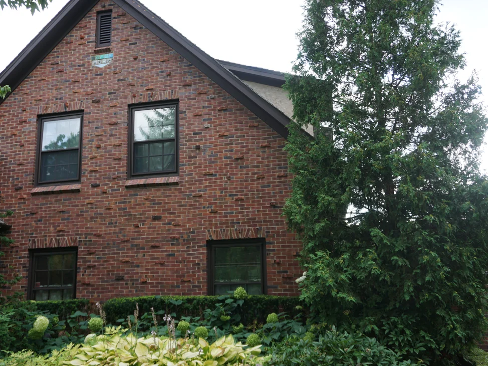 The gable end of the house in 2019, its brick a deep red brown with rough rust coloured units projecting from the face and a glazed tile panel set under the roof vent