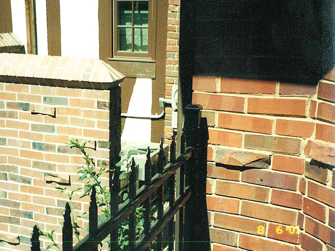 A low brick garden wall and a wrought iron gate beside the house in 2001, the wall pale and mottled next to a warmer pier with rough units projecting from it