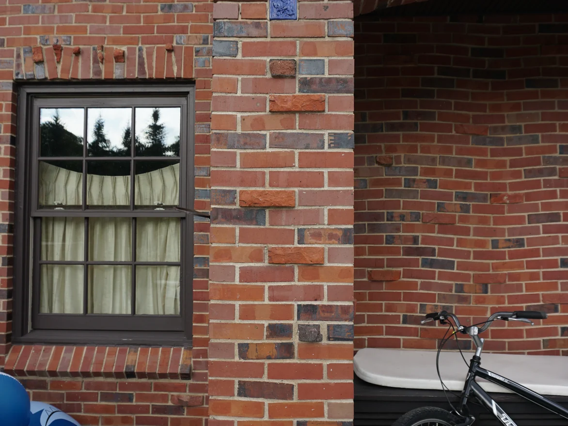 A brick pier beside a recessed arched opening in 2019, a small blue glazed tile set into the coursing at eye height and a casement window to the left