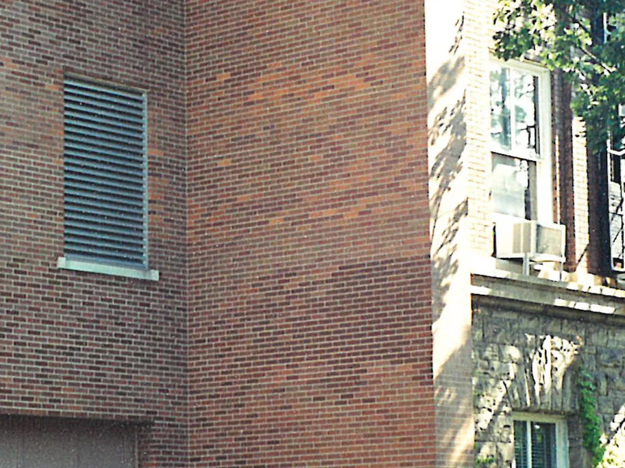 An outside corner of the university building in 2000, the brick running from pale tan to dark red in patches, with a louvered vent on the left face and an older stone-trimmed building behind