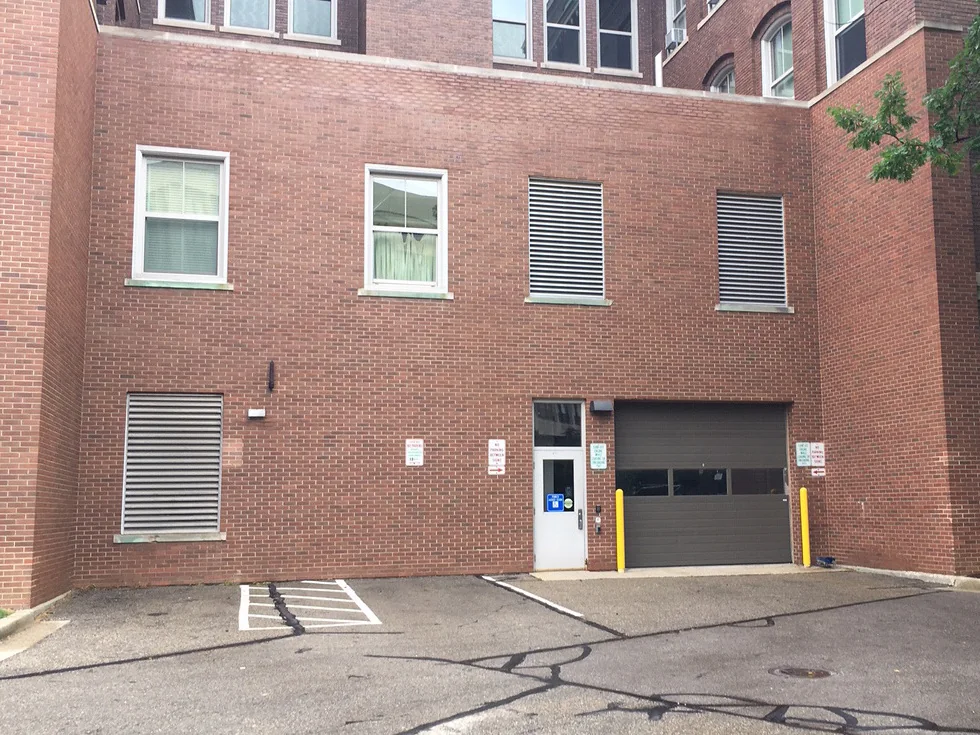 The rear elevation of the university building in 2019, two rows of windows and louvered vents in one range of red brick above a loading dock door, with the older brick building rising behind it