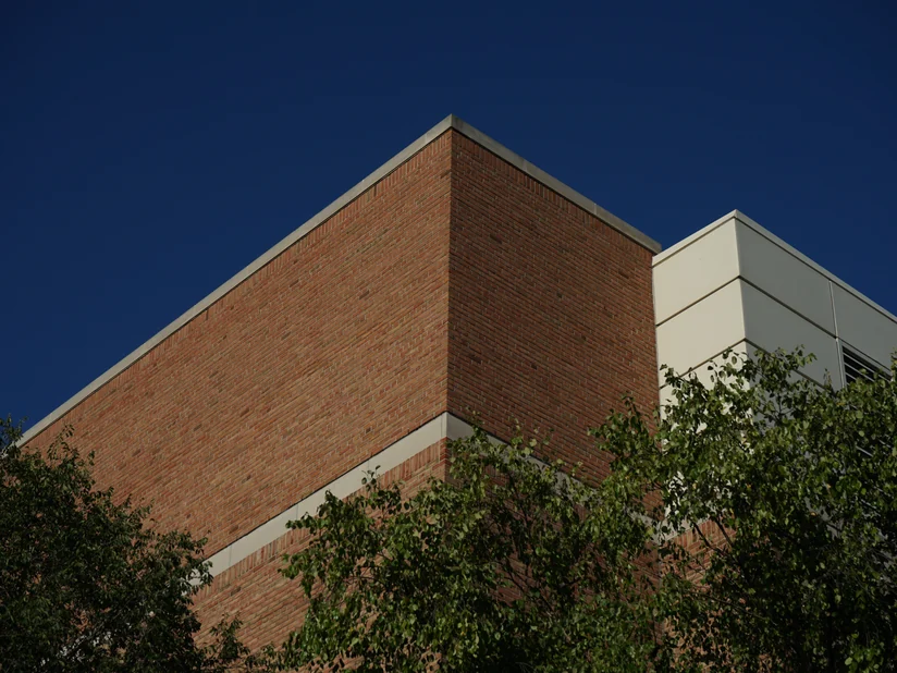 The corner of the university building in 2019, both brick elevations reading as one tan and red range up to the stone coping.