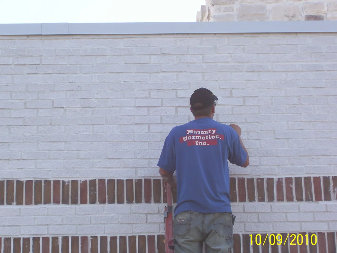 An applicator in a Masonry Cosmetics shirt on a ladder working along a white brick wall, the mortar joints in the dark brick banding below already white