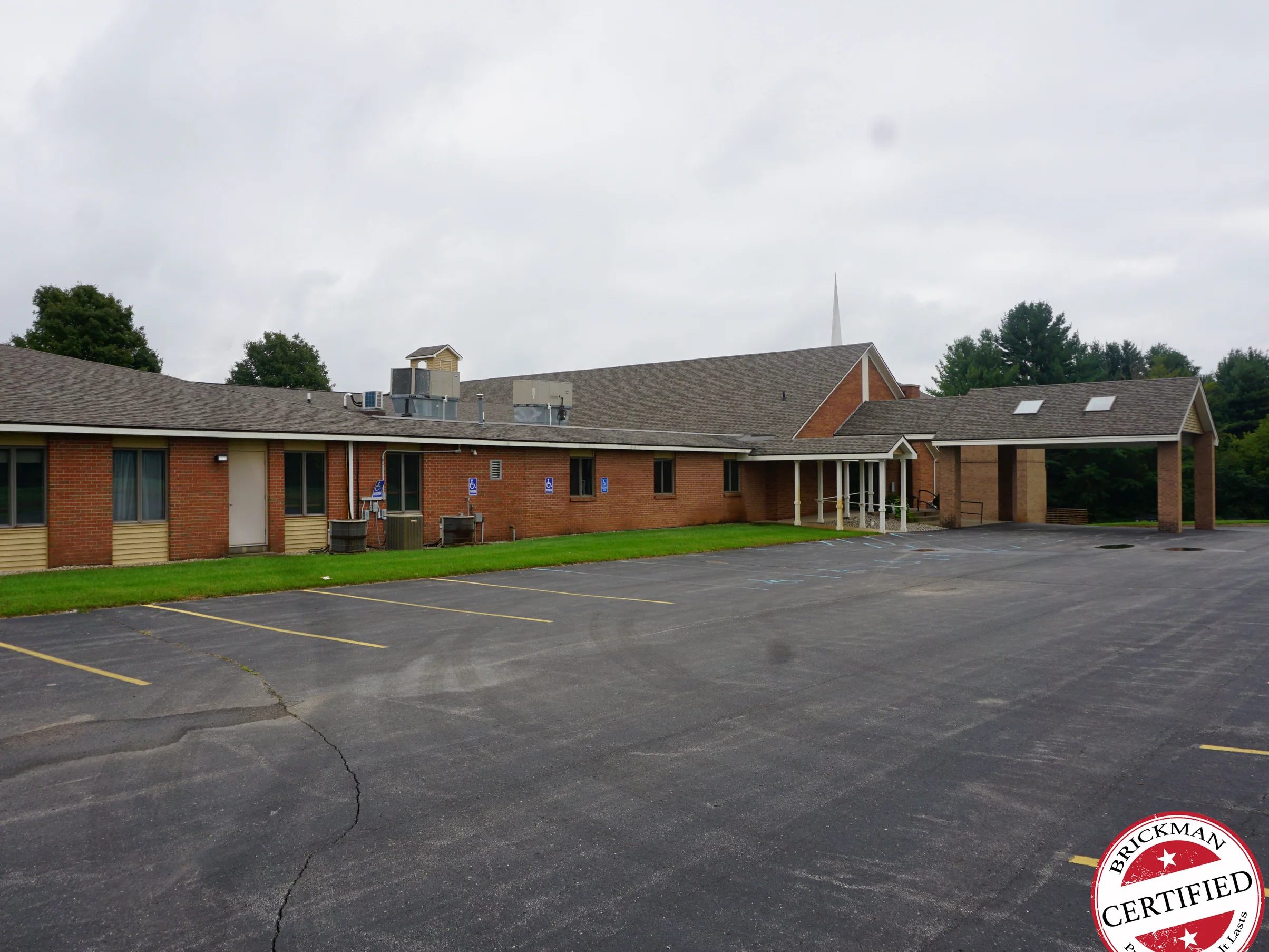 The same elevation in 2019, the long wing and the sanctuary gable reading as one even red brown under an overcast sky, with a covered drop-off at the far end