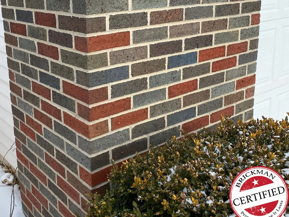 The outside corner of the brick pier at the Midland home in 2021 with snow on the ground, red and flashed blue grey units in one range beside white siding, with the certification stamp in the corner