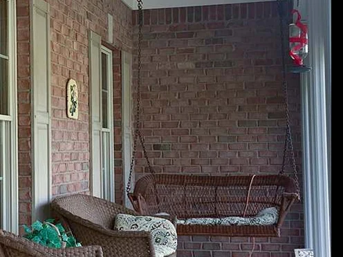 The covered porch in 2019, a wicker swing and chairs against salmon brick walls with a red lantern hanging at the far end