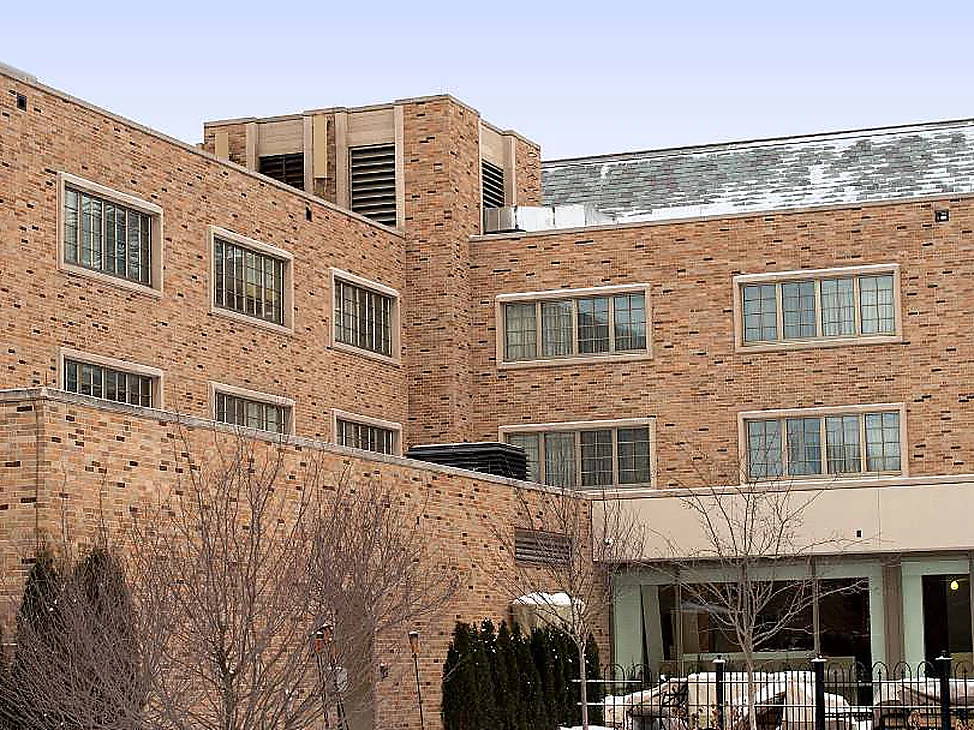 The older wing of the campus inn seen from the courtyard in 2019, three storeys of tan and brown brick over a glazed ground floor.