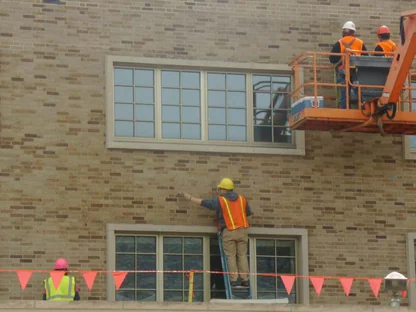 An applicator in a hard hat working stain into the brick by hand from a platform, a second crew on a boom lift at the storey above and orange safety pennants strung along the wall below
