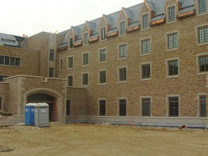 The new wing still under construction, pale buff brick running past a row of dormers to an arched entry, with bare dirt and two portable toilets in the foreground