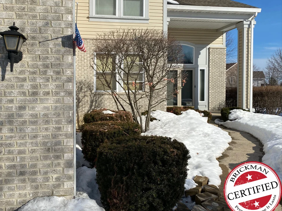 The front of the house in 2021, the grey face brick pier by the garage with its black lantern, the flag, and the covered entrance beyond, with the certification stamp in the corner
