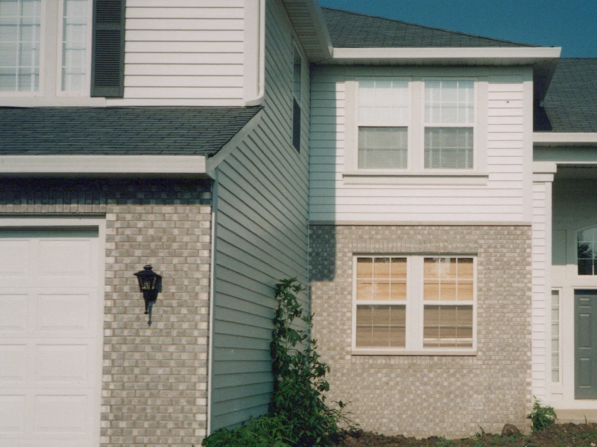 The garage corner of the house in the summer of 1998 after staining, the pier with its black lantern and the bay window base reading as one mottled grey brick, dark toothed zipper corners where the walls turn