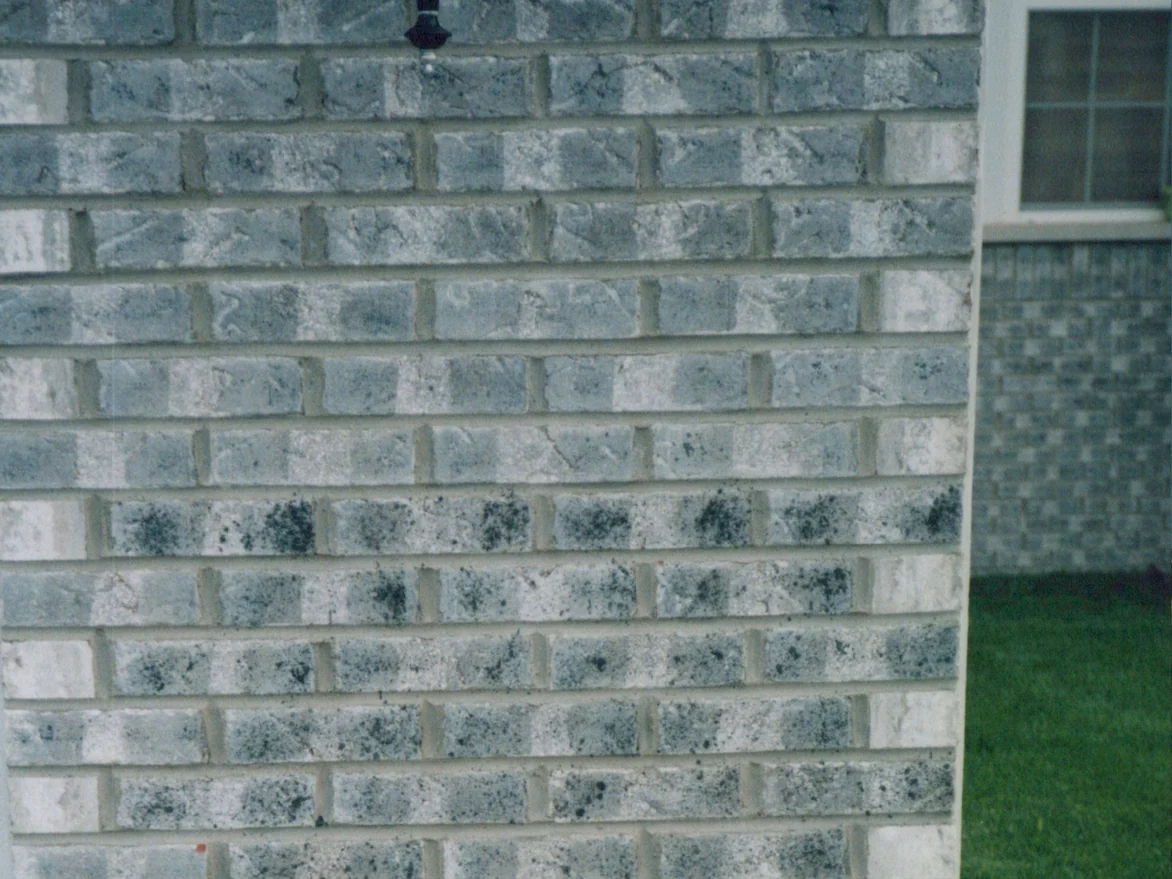 The brick pier beside the garage before staining, grey face brick with white patches across the units and a scatter of dark specks on the lower courses, the base of the lantern at the top