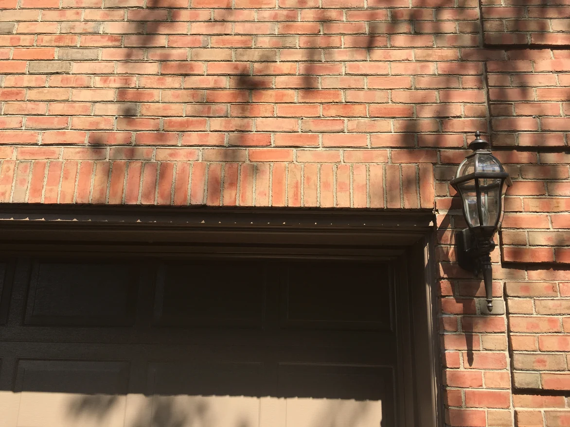 The soldier course of upright brick over a garage opening in 2019, a coach lamp on the pier beside it and tree shadow across the wall