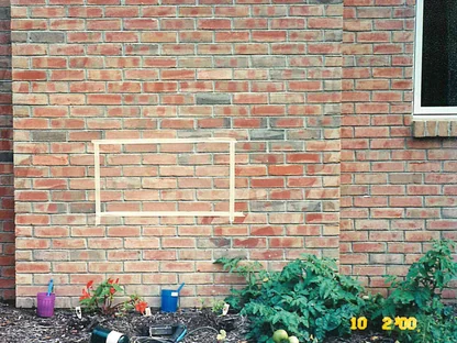 A rectangle masked off with tape on the red brick wall of a house, the wall around it blotched pale where it had been washed