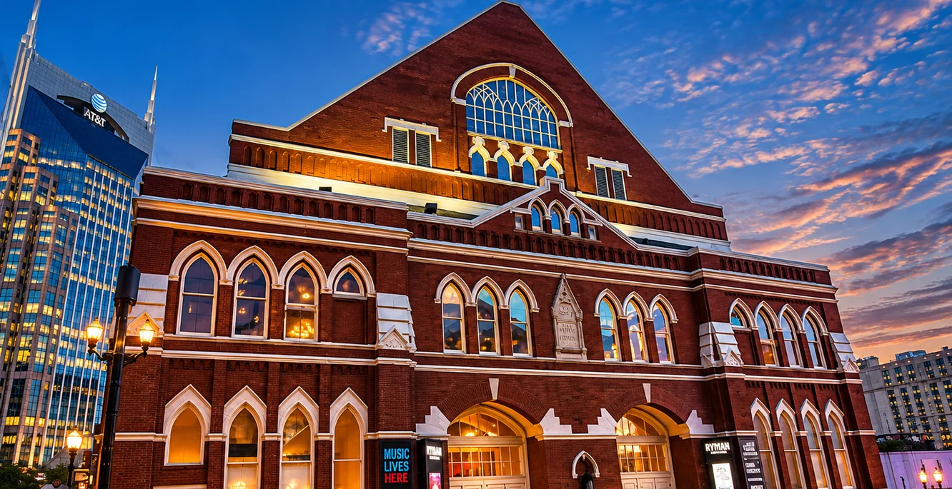 The Ryman Auditorium in downtown Nashville at dusk, its red brick gable and arched windows lit from inside