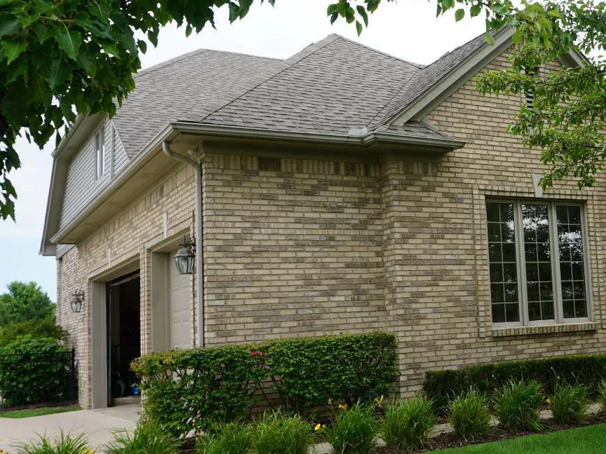 The garage-side corner of the house in 2019 under a mature tree, the brick one even tan and buff blend from the garage door past the tall window to the gable