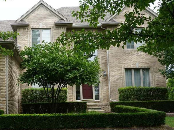 The entry gables in 2019 behind a small tree and clipped hedges, the brick around the dark red front door and the upper windows holding one range