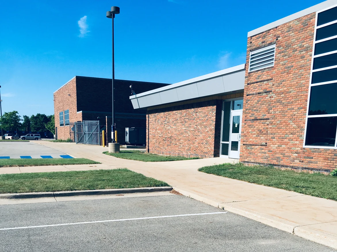 A glass entrance door set into the school's brick corner in 2019, with a second brick block and a fenced service yard behind it