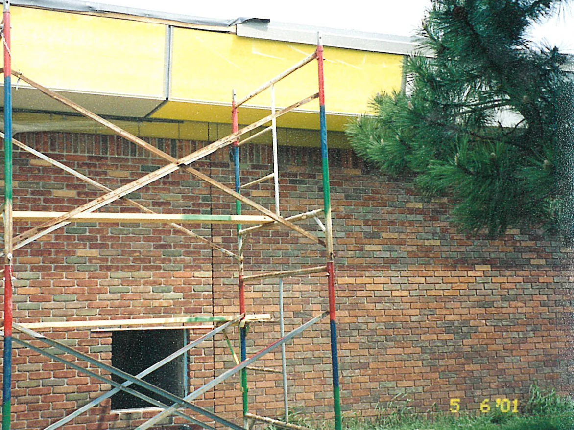 Scaffolding standing against a low school wall in 2001, the brick behind it in mixed reds, tans and greens under a yellow fascia