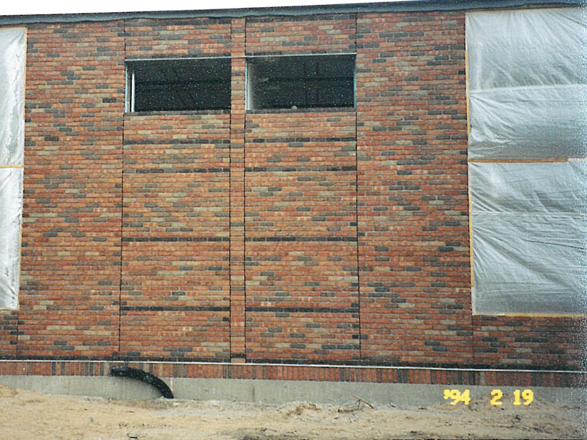 A panel of school wall still under construction, sheeting over the openings and bare ground at its base, the brick running in mixed reds, tans and near-blacks with darker courses across it