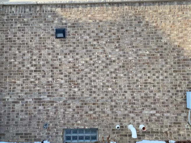 The side wall in 2021, the brick even across the courses above a glass block basement window and two vent pipes, with a light fixture on the field