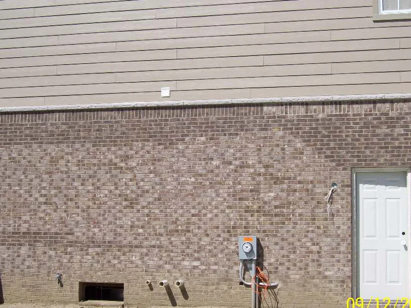 The garage side wall in September 2007, an electric meter and a white service door on brick blotched light and dark, with cut lumber and pipe lying on bare dirt