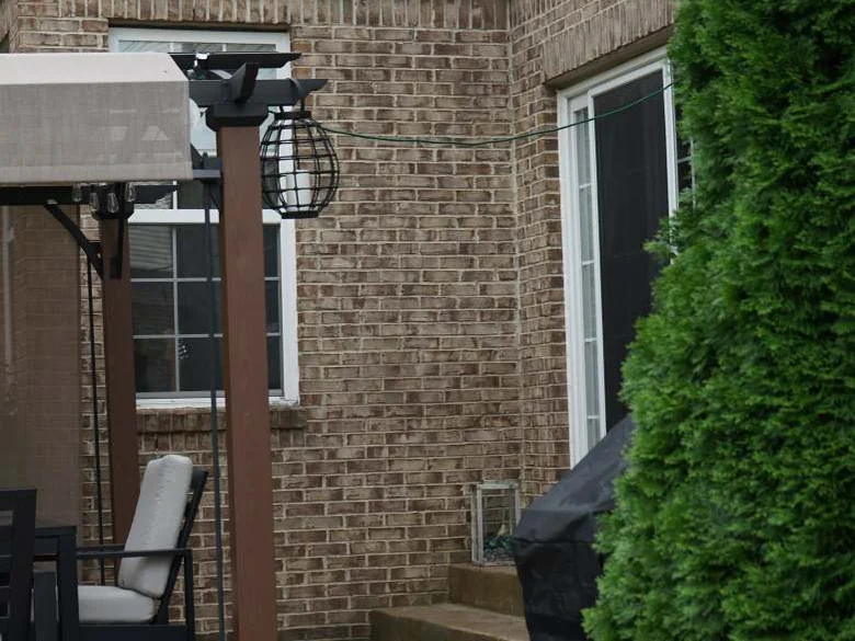 The rear patio corner in 2019, a pergola post and lantern in front of a brick wall with a tall window and an evergreen beside it