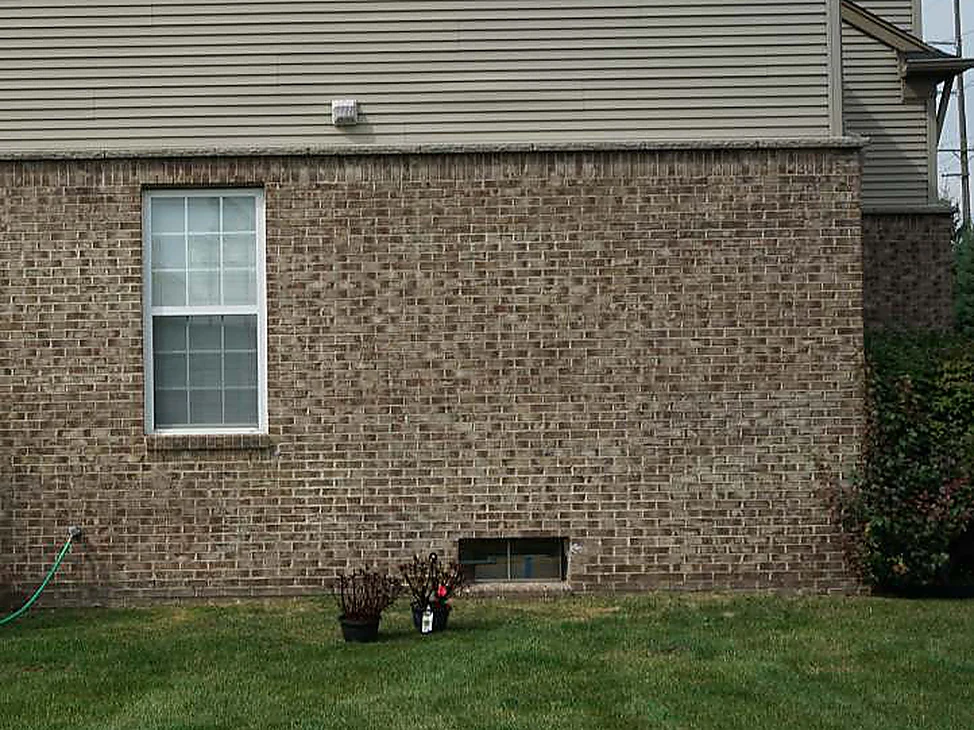 The side wall of the Northville house in 2019, tan brick under vinyl siding reading as one range from the window down to the lawn