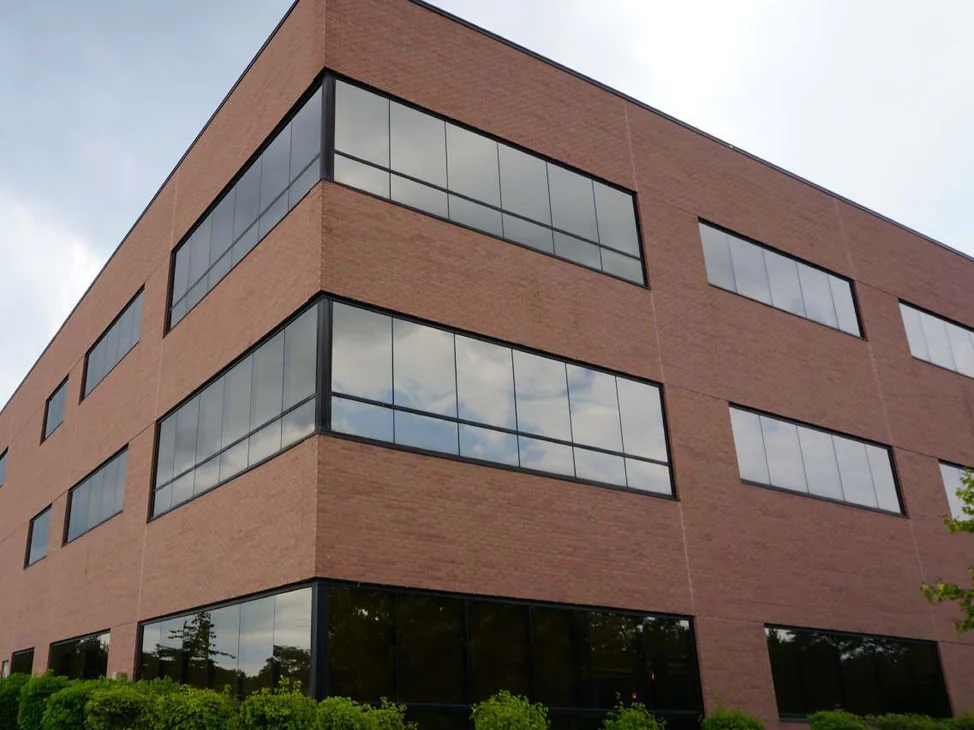 The outside corner of the three-story office in 2019, red brick running past three rows of ribbon windows above a clipped hedge, with an overcast sky behind