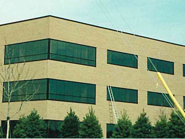 The corner of the office in 1999 with a yellow boom and an extension ladder against the tan brick wall and young pines along the base