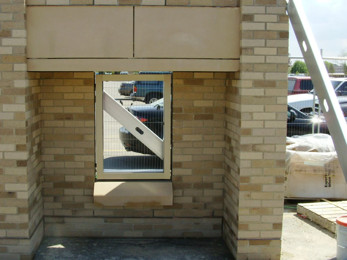 A freestanding brick mockup panel built in a car park with a window and cast stone sill, its brick running pale and even with almost no dark units