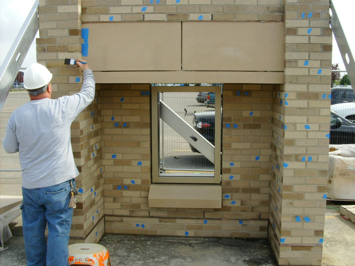 An applicator in a hard hat brushing stain onto single brick units on the same mockup panel, with blue tape marking every unit that is to be darkened