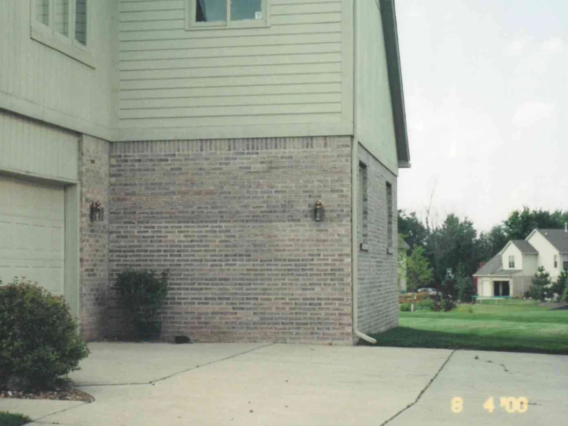 The corner of the house beside the garage door in August 2000, grey and tan brick under a soldier course below pale green siding, with a shrub against the wall and the camera date in the corner