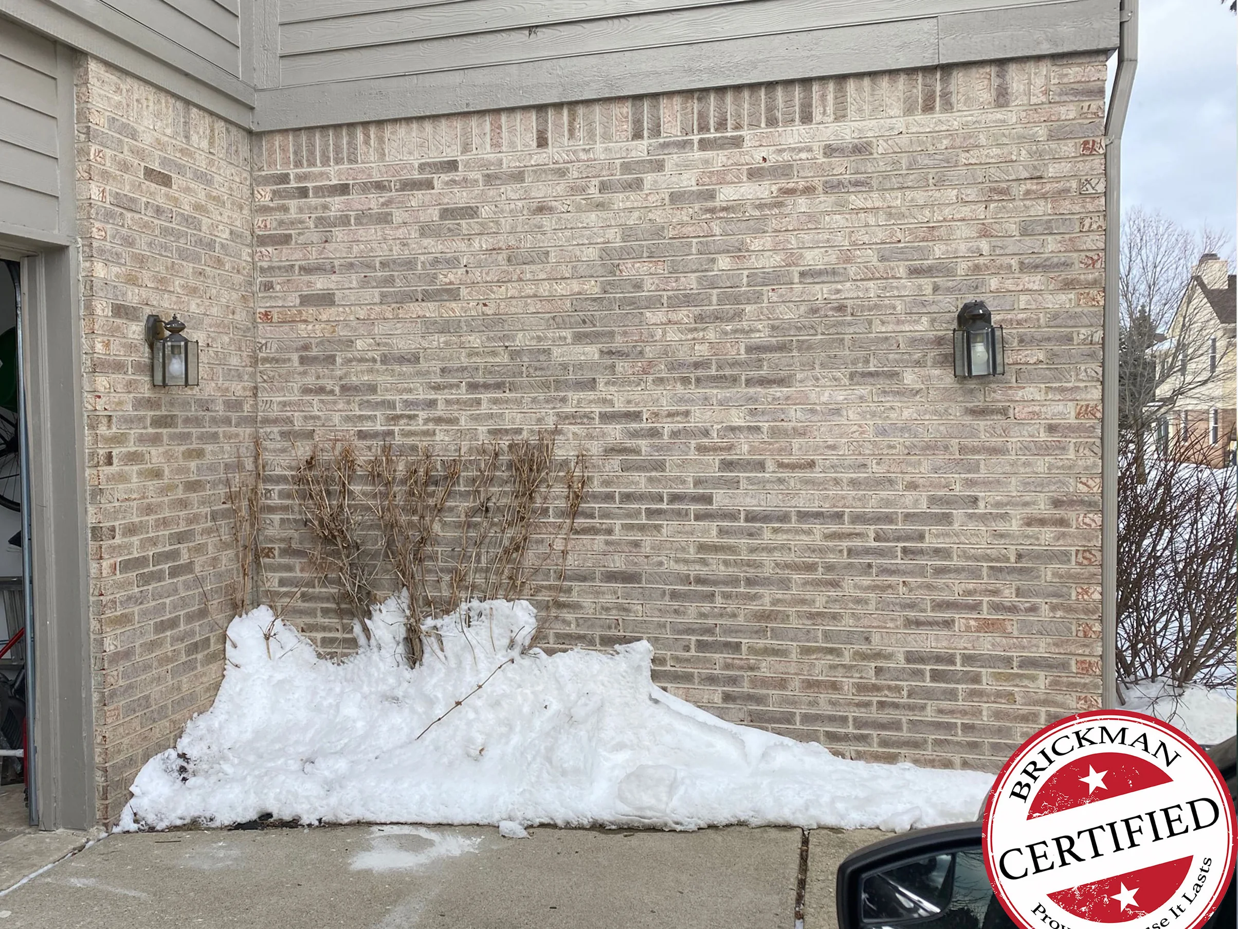 The garage corner of the house in 2021 with snow banked against the wall, the brick one pale range up to the tan siding, with the certification stamp in the corner