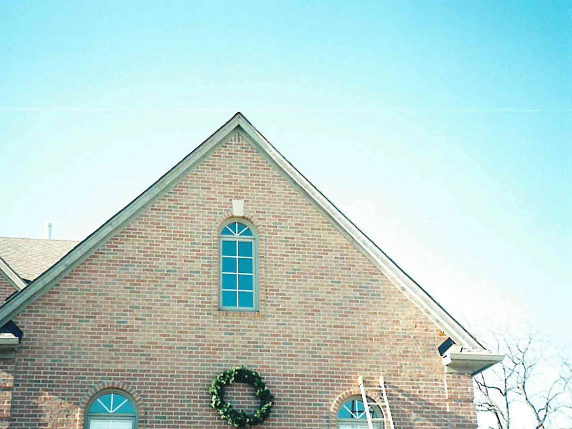 The main gable of the house in 1998 under a bare winter sky, the brick running pale tan and pink with darker units gathered low on the wall, a wreath hung below the arched window