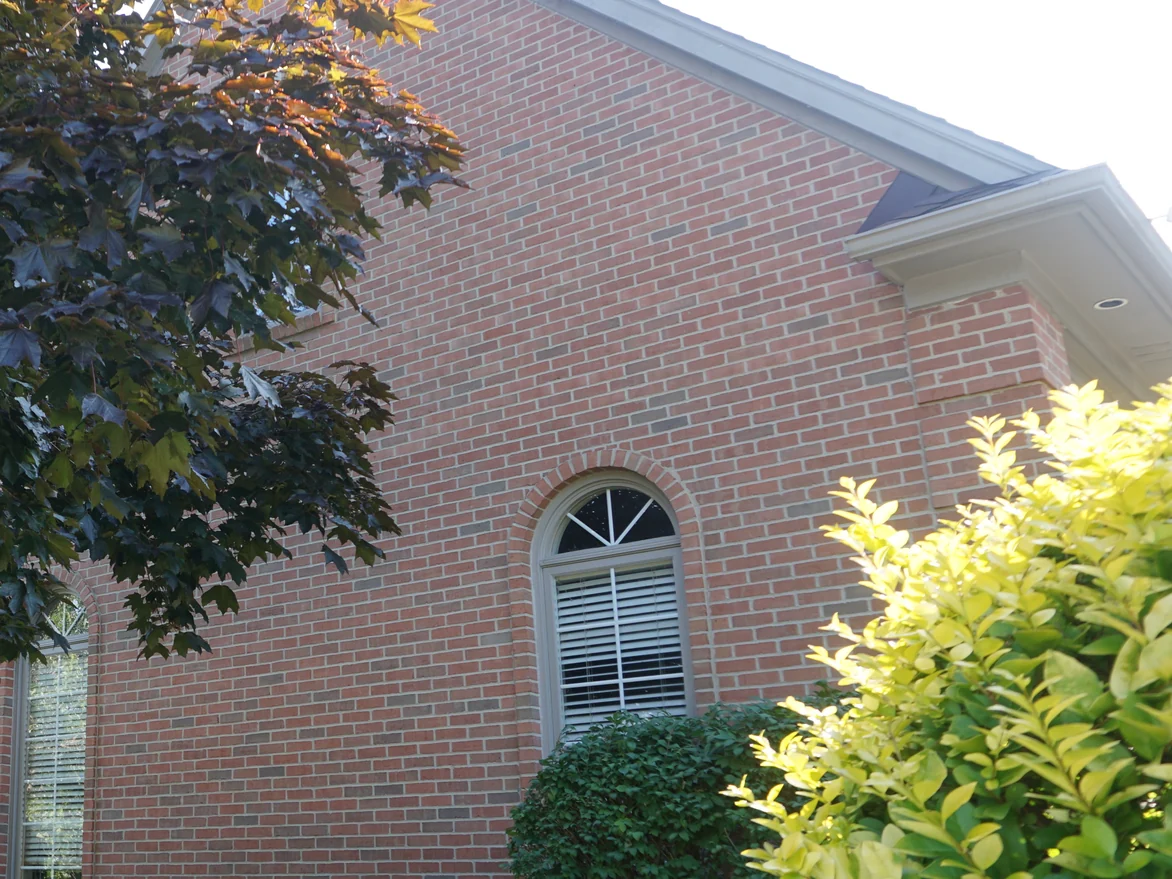 The gable end of the house in 2019 seen past a maple, the wall one range of red and tan with darker headers scattered through the courses