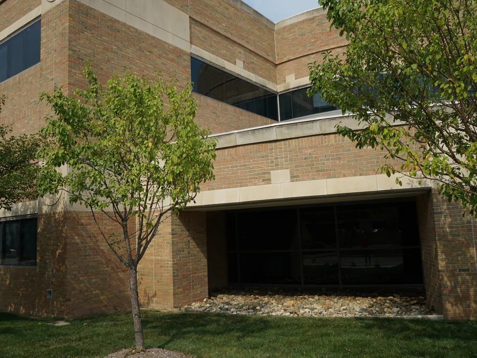 An outside corner of the hospital in 2019 beside a recessed glazed bay and a bed of river stone, the brick holding one range from the ground to the coping