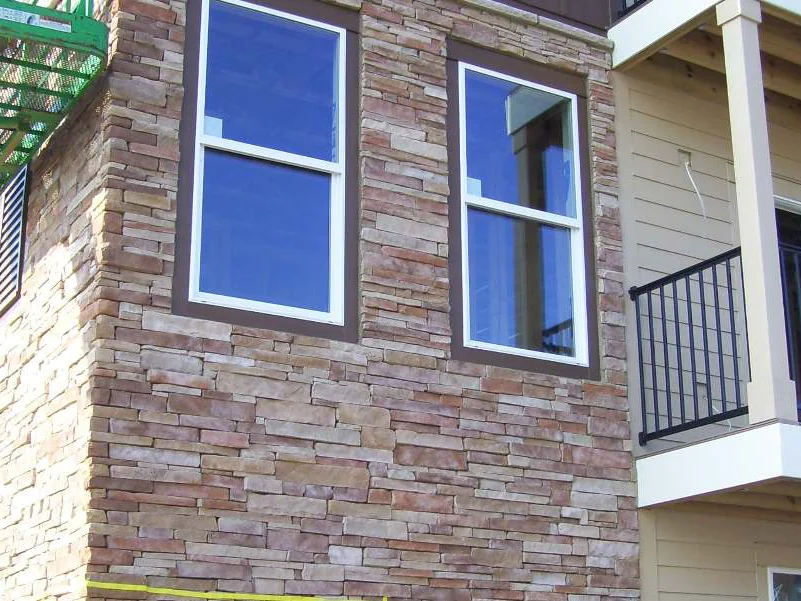 An outside corner beside a balcony in 2009, two windows set in stacked stone that runs cream through pale pink, with a green boom lift at the top left