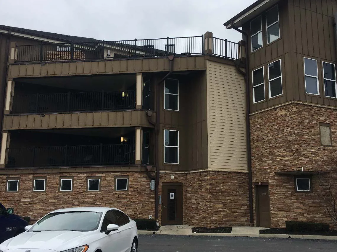 The apartment building from the parking lot in 2021, three storeys of balconies and board siding over a continuous stone base course, a white car in the nearest bay