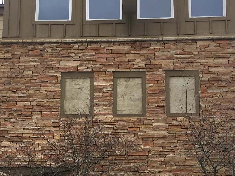 The same wall in 2021 under an overcast sky, the three openings now flat panels rather than louvers, the stone one blended range of red brown and tan with bare saplings in front