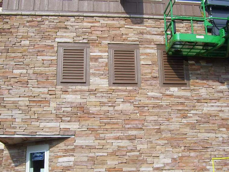 A stone wall with three brown louvered vents in 2009, the stone reading cream and pale tan in full sun, a green boom lift parked against the upper right corner