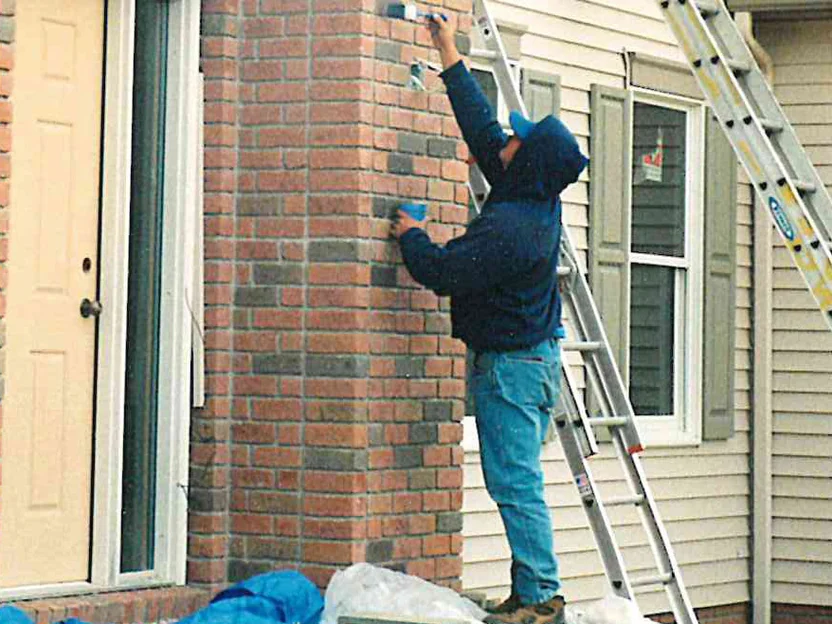 An applicator standing on the front porch in 1998, working stain into the brick corner by hand with ladders leaning on the siding beside him