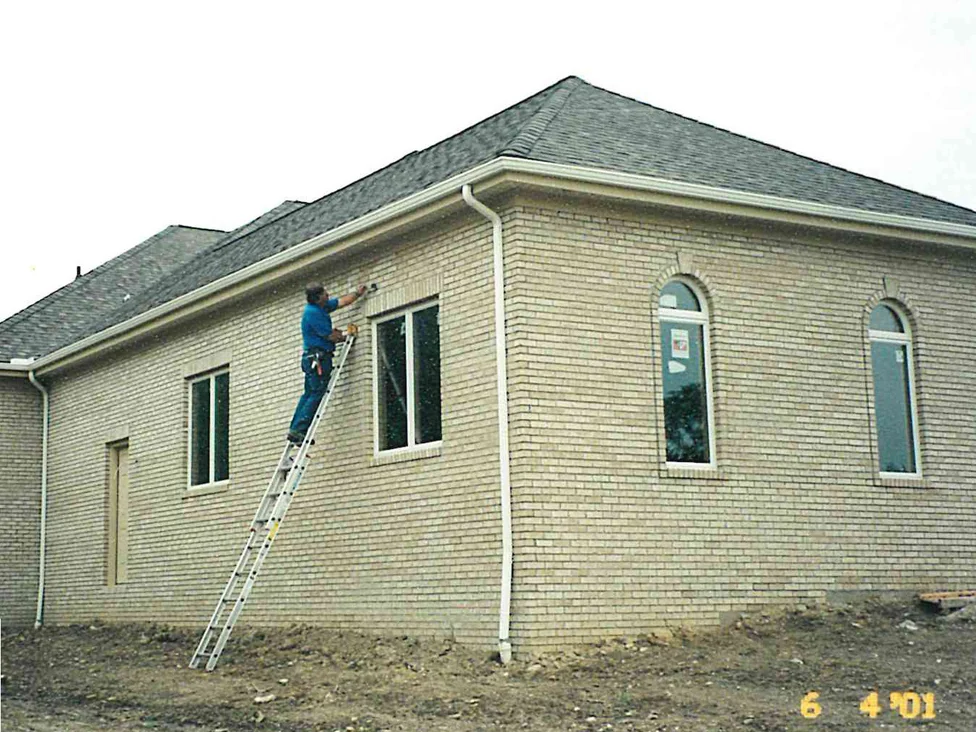 A worker on an extension ladder brushing stain onto the cream brick side wall of the single-story house in 2001, the hipped roof running back over bare graded dirt and two arched windows on the end wall to the right