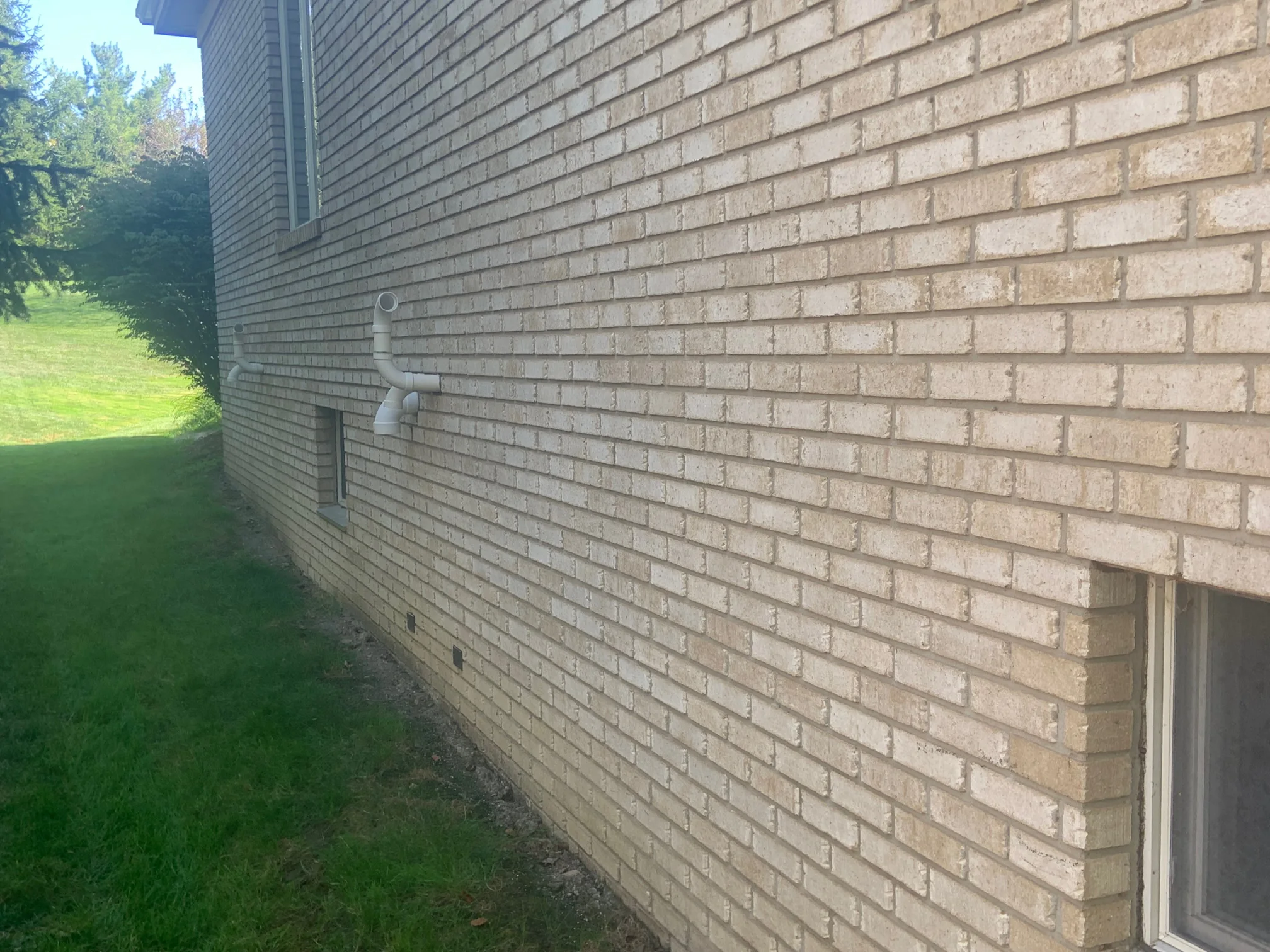 The long side wall of the house in 2019 seen at a raking angle, the stained brick running away toward the roofline with mown lawn and evergreens beside it