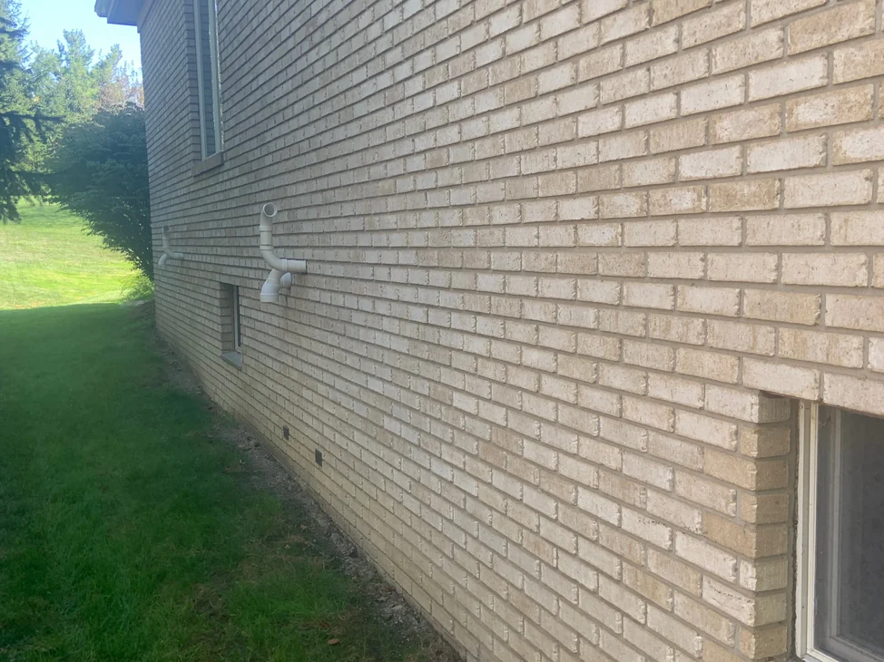 The long side wall of the house in 2019 seen at a raking angle, the stained brick running away toward the roofline with mown lawn and evergreens beside it.