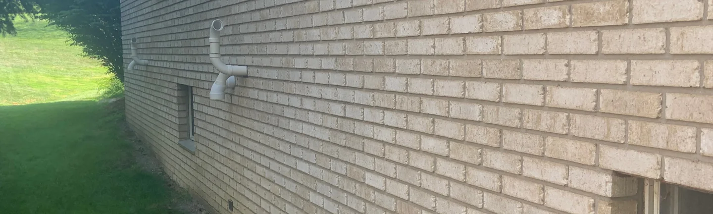 The long side wall of the house in 2019 seen at a raking angle, the stained brick running away toward the roofline with mown lawn and evergreens beside it