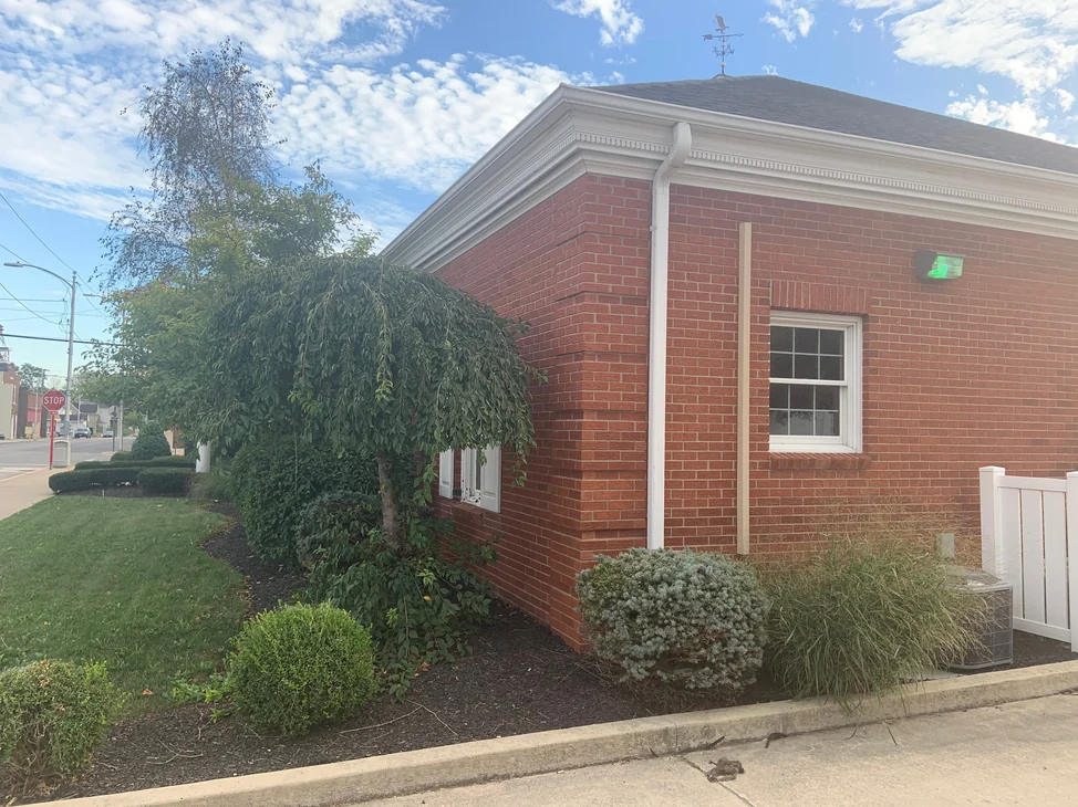 The outside corner of the bank in 2019, red brick running past a quoined corner to a white cornice, with a weeping tree and planted beds along the lawn