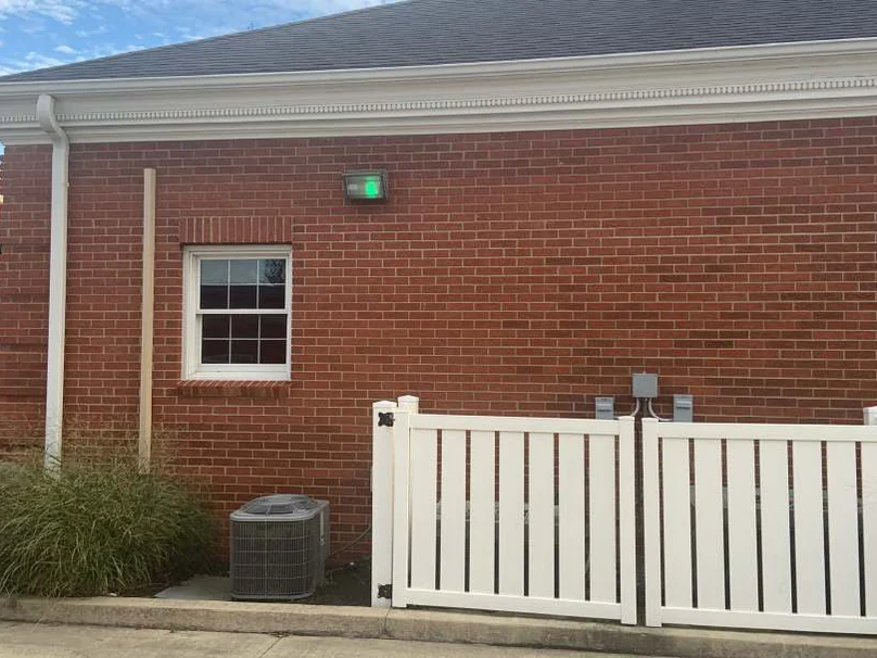 A rear wall in 2019, red brick around a white window with a white fenced equipment enclosure and an air conditioning unit at the base