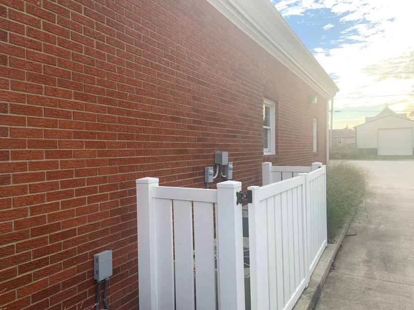 A raking view along the long red brick wall in 2019, running past meter boxes to a white vinyl gate and the drive beyond